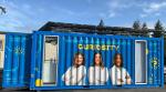 lays three The Curiosity Cube, a blue mobile science lab that displays three students in white lab coats and text that reads "Breakthroughs Begin With Curiosity"